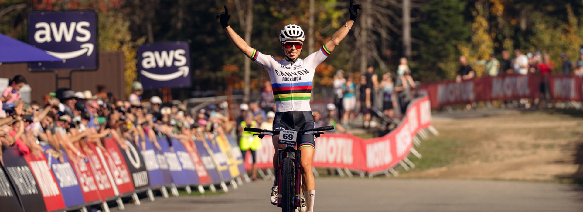 A female mountain biker crosses the finish line at Mt Van Hoevenberg with her hands raised triumphantly in the air.