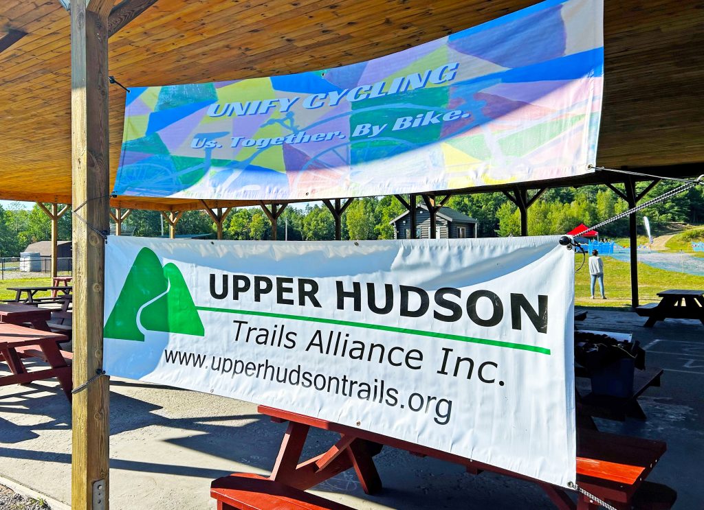 "Unify Cycling" and "Upper Hudson Trails Alliance Inc." banners hang in the shelter near the start line. 