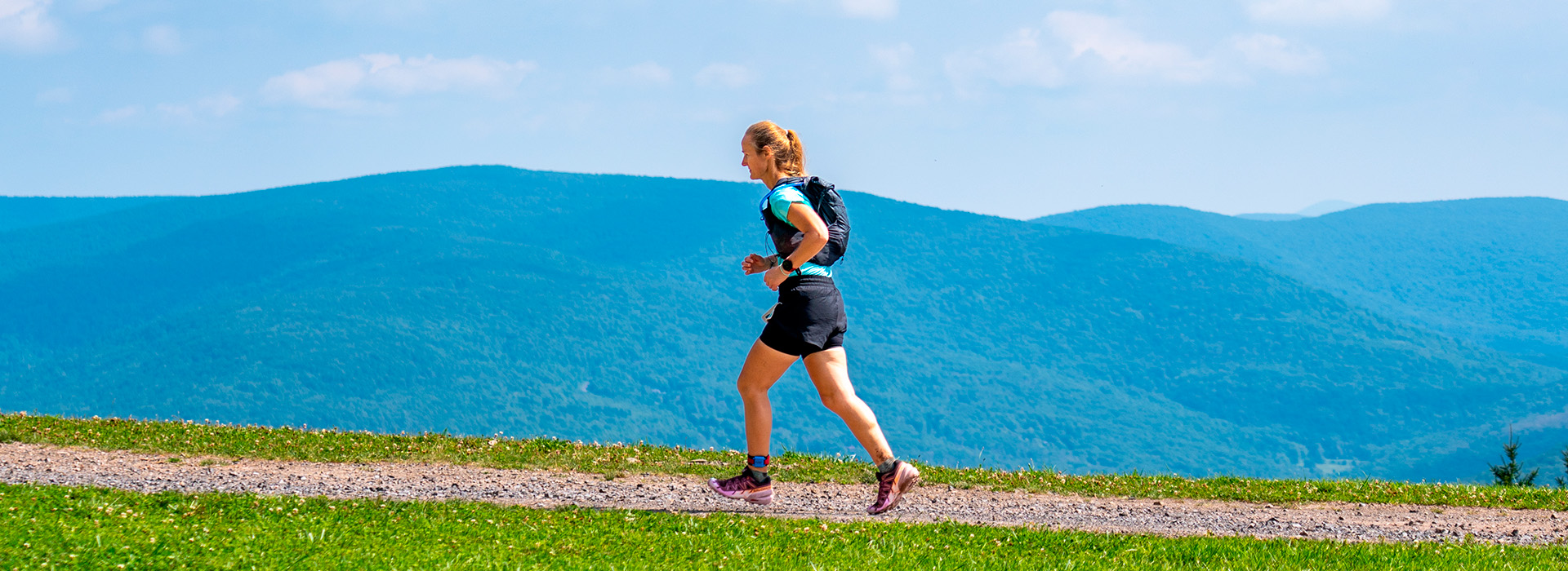 A woman with a small backpack runs across the top of Belleayre Mountain during the Climb It for Climate race with a beautiful view of the Catskills in the background.
