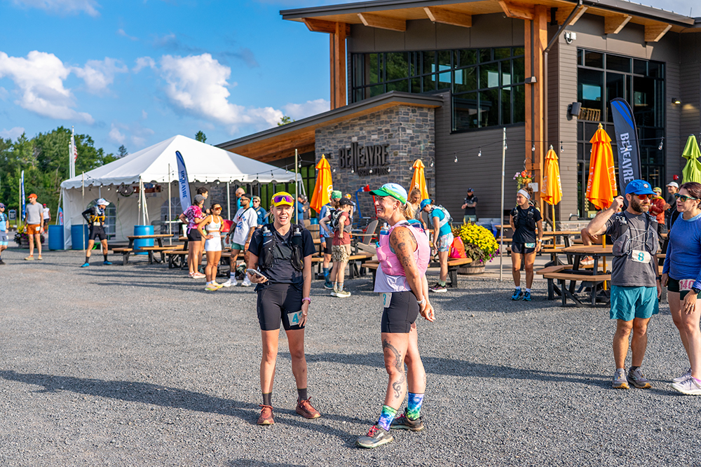 Two women runners stand outside the Belleayre Discovery Lodge talking prior to beginning the trail race. Others runners and the lodge are in the background.