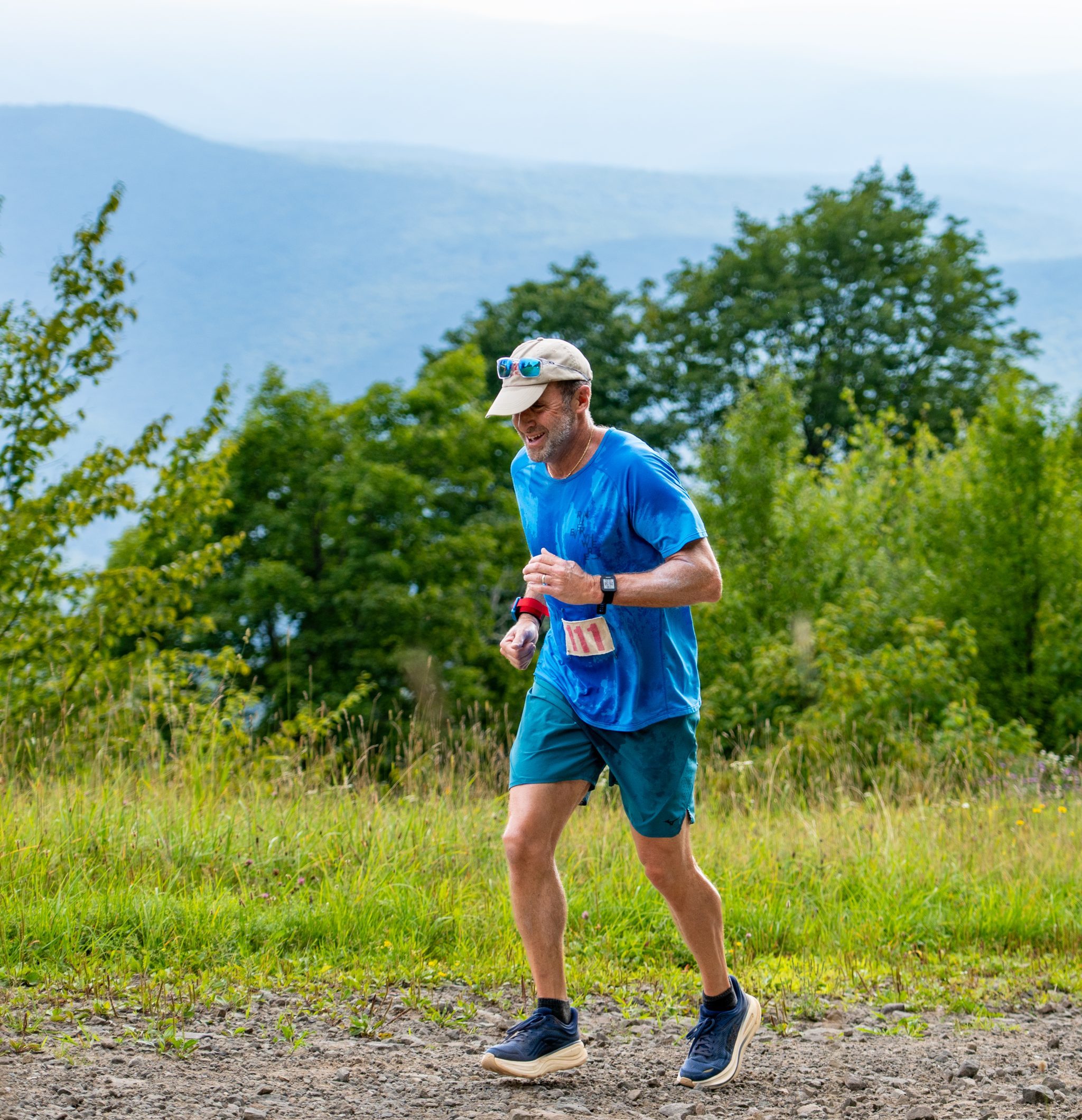 A male runner in blue shorts and a blue t-shirt runs upward on a ski trail on Belleayre Mountain with a view of the Catskills behind him.