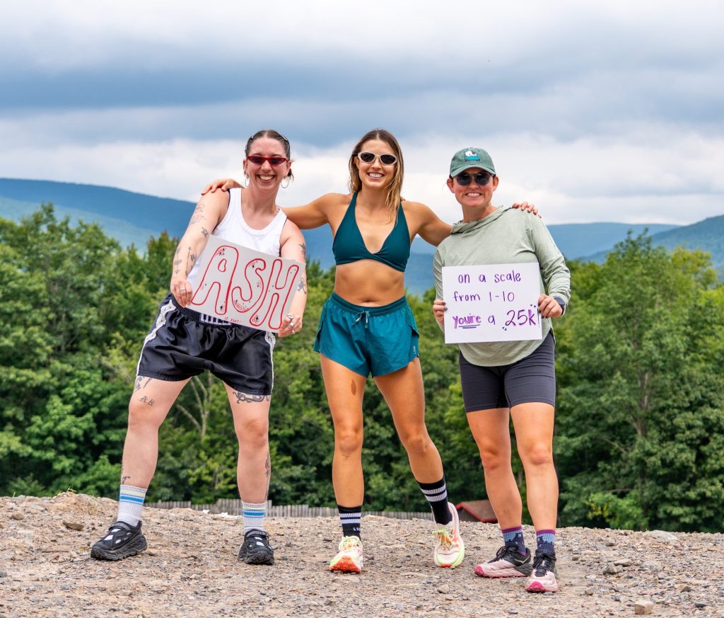 Three women atop Belleayre Mountain stand together smiling with signs that read "ASH" and "on a scale from 1-10, you're a 25k."