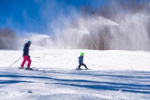 A child with an adult skiing down a slope at Belleayre mountain with high efficiency snowguns blowing snow in the background. 