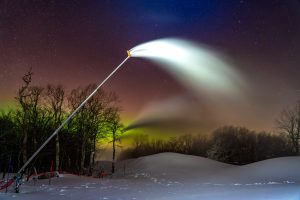 Nighttime at Belleayre Mountain with a high-efficiency snow gun operating in the foreground and the Northern Lights glowing in the forest in the background. 