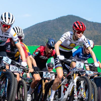A mass start of a women's UCI mountain bike race close up with many riders and a mountain and blue sky in the background.