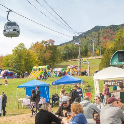 People enjoy Oktoberfest at Whiteface whiile the gondola goes overhead against the backdrop of fall leaves on the mountain.