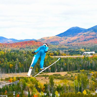 A ski jumper in a blue suit sails through the air over a colorful fall landscape with mountains in the background.