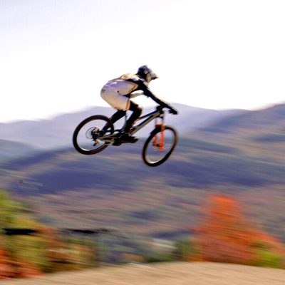 An elite mountain bike rider sails through the air high off a jump at Whiteface Mountain with fall foliage and mountains in the background.