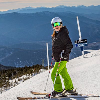 A female skier stands alone with a big smile on top of the Skyward run far up on the mountain in winter with a beautiful view of the mountains and landscape below.