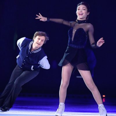 A female figure skater smiles brightly as her male pairs counterpart skates around her and under her outstreached arm.