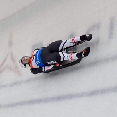 A male luge athlete slides down the track across a big tall turn with the words "Lake Placid" visible in the ice below.