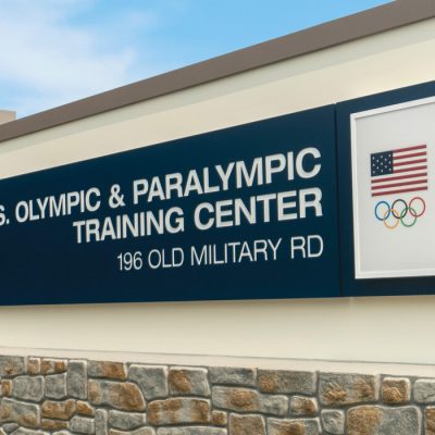 Image of the large sign for the U.S. Olympic & Paralympic Training Center, 196 Old Military Road with two american flags and Olympic symbols.