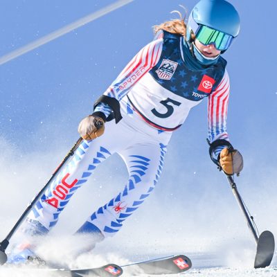 Kelsey O'Driscoll skis with outriggers wearing a white Team USA uniform, all against a blue sky.