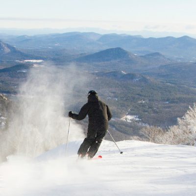 Solo skier in black at the top of a run on a winter day with a view of the mountains in the background.