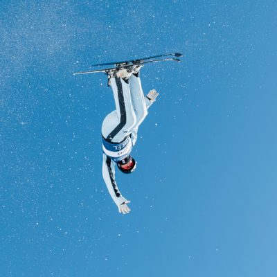 An aerials athlete upside down in the air while performing a trick high in the sky against the blue background.