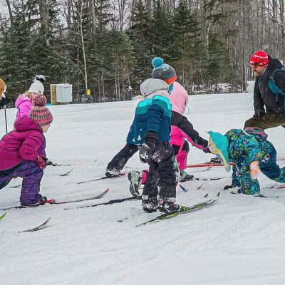 A group of young skiers dressed in winter gear practicing a move on skis their instructor is demonstrating for them.