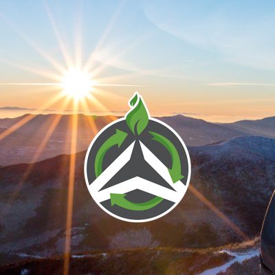 A winter sunrise view from the summit of Whiteface with a skier on the right and the green and white Olympic Authority sustainability logo in the center with the sun shining on it.