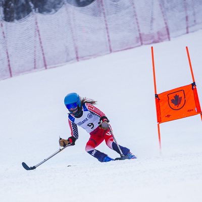 Kelsey, in a red ski racing leotard, speeds down a slope, passing an orange gate with safety fencing and trees in the background.