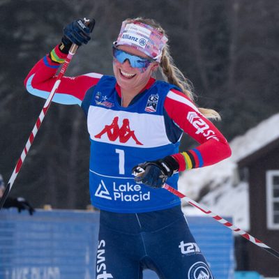 Cross-country skier Jessie Diggins smiles and raises her arm in the air triumphantly as she crosses the finish line at a Mt Van Hoevenberg race.