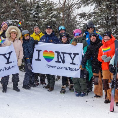 A large group of skiers on the mountain in the snow with trees in the background, some of them holding two white banners with the I Love NY logo, including a rainbow heart in each.