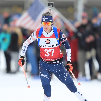 A female biathlete skis hard to the finish line at Mt Van Hoevenberg with blurred spectators in the image behind her.