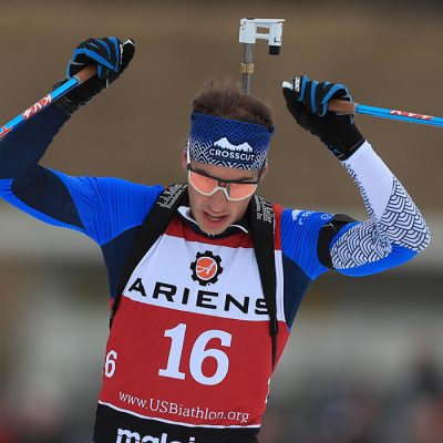 A male biathlete raises his hands in triumph as he crosses the finish line.