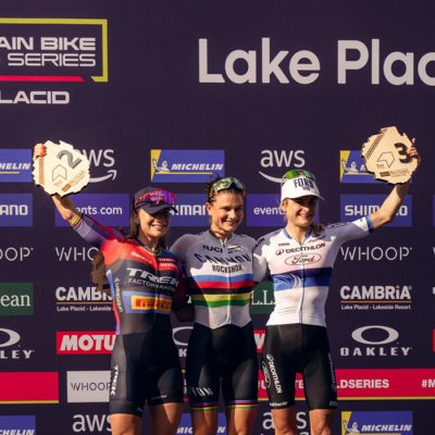 Three women stand triumphantly and holding trophies on the podium after their race at Mt Van Hoevenberg in Lake Placid with the UCI World Series and Lake Placid logos in the background.