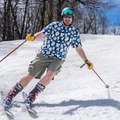 A man in a Hawaiian shirt and shorts skis a snowy slope with trees and blue sky behind him.
