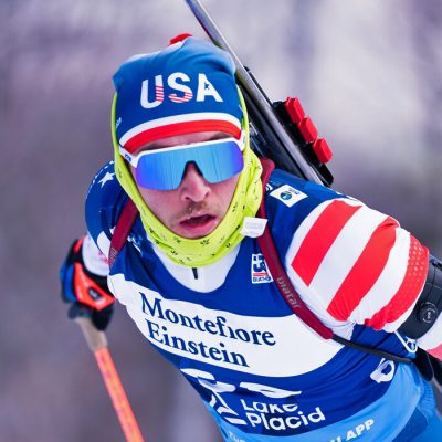 An IBU Cup athlete bearing a USA hat, looks hard through his sunglasses as he works hard skiing in competition on a winter day.