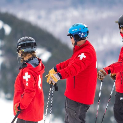 Four ski patrol members of the Whiteface Mountain crew, all in red Mammut jackets on the summit looking down on Little Wihteface.