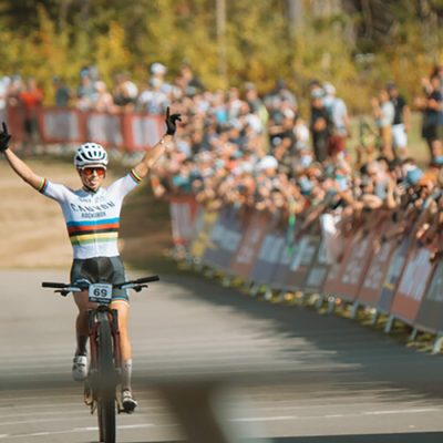 A female rider in a UCI mountain bike cross country race raises her arms in triumph as he crosses the finish line in front of many spectators in an outdoor fall scene.