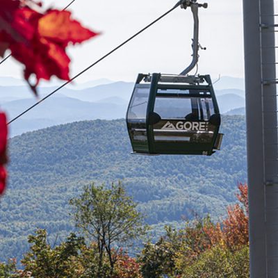 A cabin of the Northwoods Gondola rises high on Gore Mountain with fall foliage in the foreground and mountains in the background.