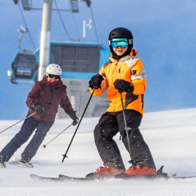 A family of four skis down a slope facing the camera with the gondola overhead and blue sky behind them.