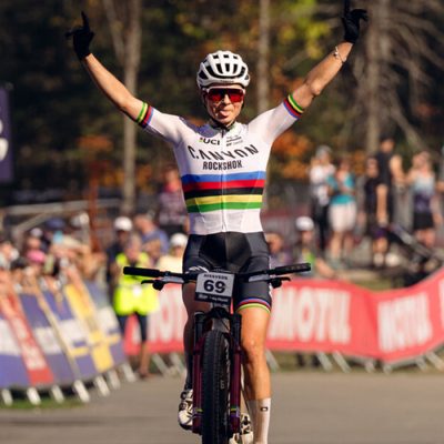 A female mountain biker crosses the finish line at Mt Van Hoevenberg with her hands raised triumphantly in the air.