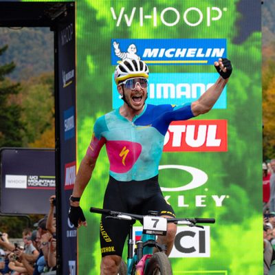 A male MTB rider finishes a race triumphantly with his fist in the air with spectators and the finish arch and sponsor list in the background.