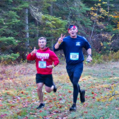 Two young men trail running in the fall at Mt Van Hoevenberg with one giving a thumbs up and smiling and the other showing a peace sign.