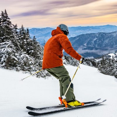 A skier in an orange outer layer seen from the side skies down a slope high on Whiteface Mountain with slightly orange clouds in the background and trees surrounding the slope.