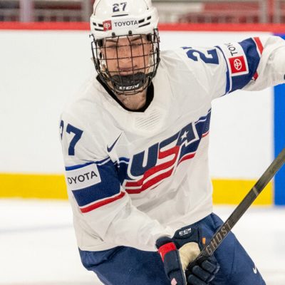A female hockey player in a helmet and her USA jersey handles a puck on the ice.