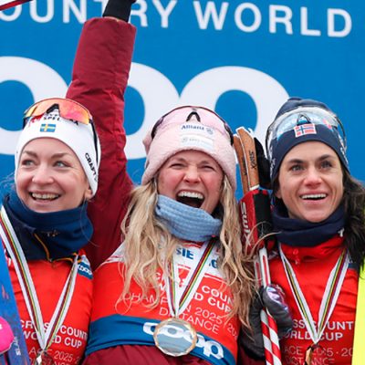 U,S. Cross-country ski legend Jessie Diggins on the podium between two other competitors raising her skies triumphantly in her left arm while smiling and cheering.