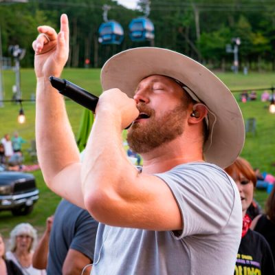 Close up of a male singer with a microphone and wearing a brown wide brim hat pointing to the sky with dancers and other people gathered nearby and the gondola and grass and trees in the background.