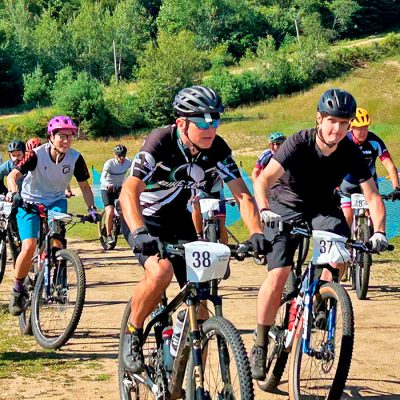 A group of mountain bike riders starts off a 6 hour race at the North Creek Ski Bowl with green trees in the background.