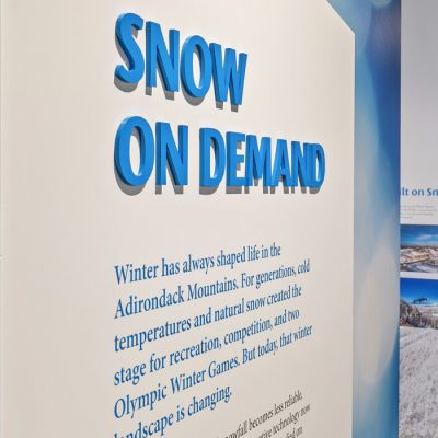 Close up of the first panel of the Snow on Demand exhibit in the Olympic Museum with the title of the exhibit in the foreground and a large image of snowy ski trails in the background.