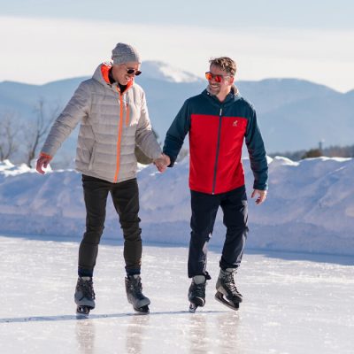 Two men skate together on the Olympic speed skating oval, one helping the other by holding his hand, with the Adirondack Mountains in the background.