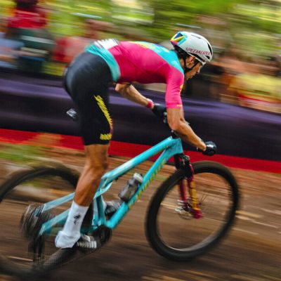 A male mountain bike rider goes fast through the forest in the UCI Cross-Country World Cup with blurred spectators in the background.