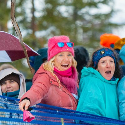 Spectators line a cross country ski course cheering happily with the legs of a US Ski Team competitor coming through in the foreground.