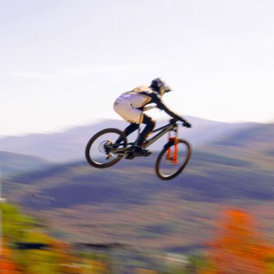 A downhill mountain bike racer flies high over a jump at Whiteface Mountain with the Adirondack landscape and fall foliage in the background.