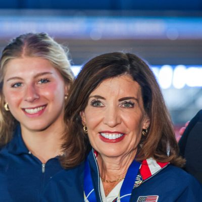 Governor Hochul smiling inside the 1980 Herb Brooks Arena in Lake Placid side by side with two gold medal winning women hockey players and one of their coaches.